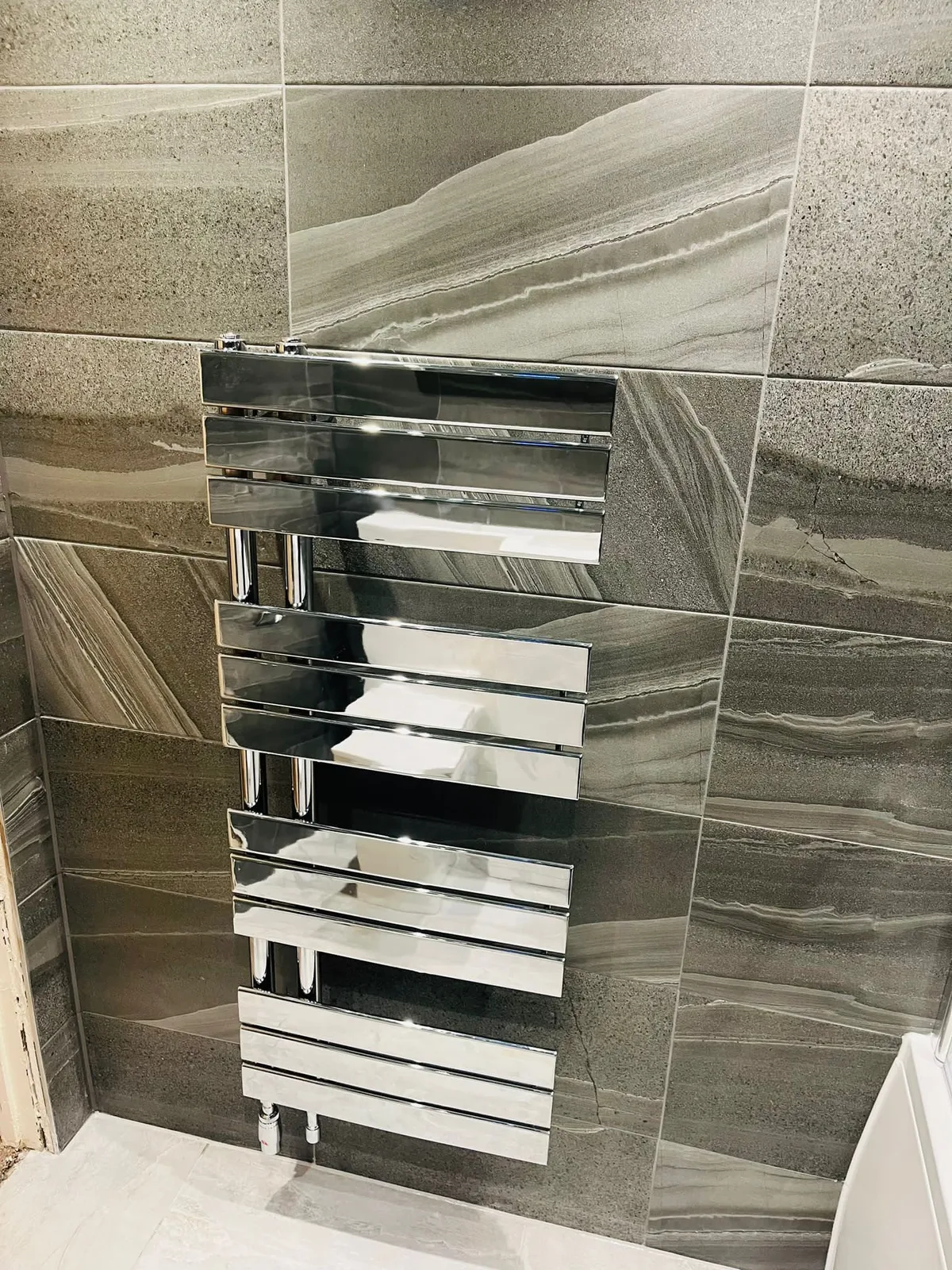 Chrome heated towel radiator fitted to a grey tiled bathroom wall